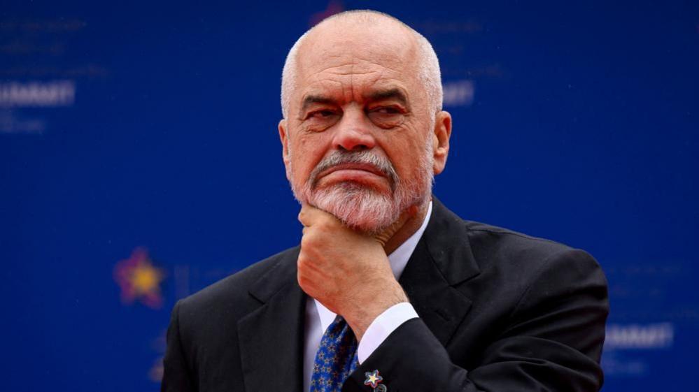 A man with a grey beard and moustache puts his hand on his chin and frowns