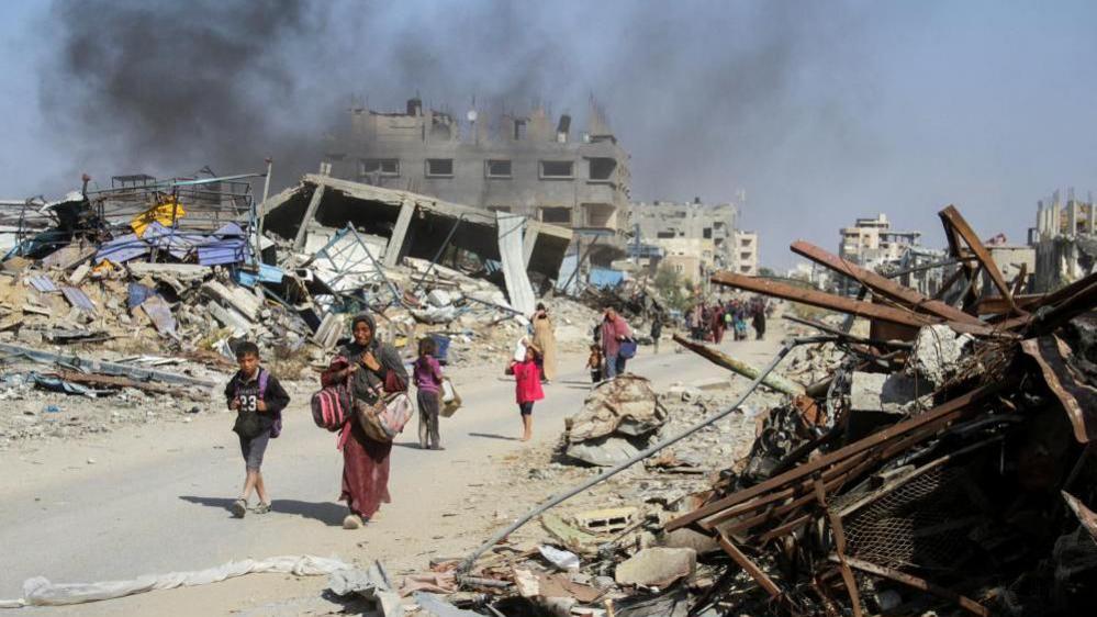 women and children walk past the smouldering wreckage of buildings in Gaza