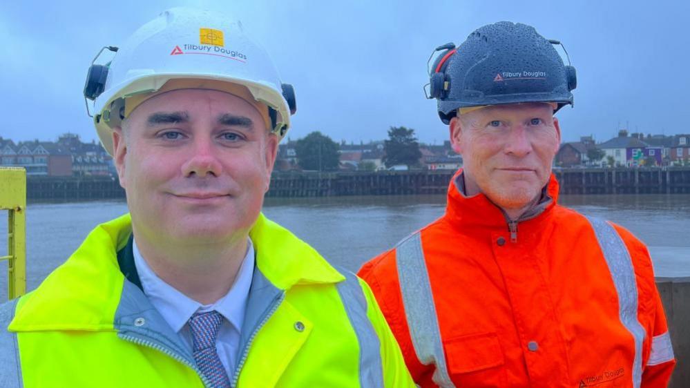 Daniel Candon, left in yellow high visibility jacket and Steve Kirk, right, in orange high visibility jacket. Both are wearing hard hats, standing near the River Yare