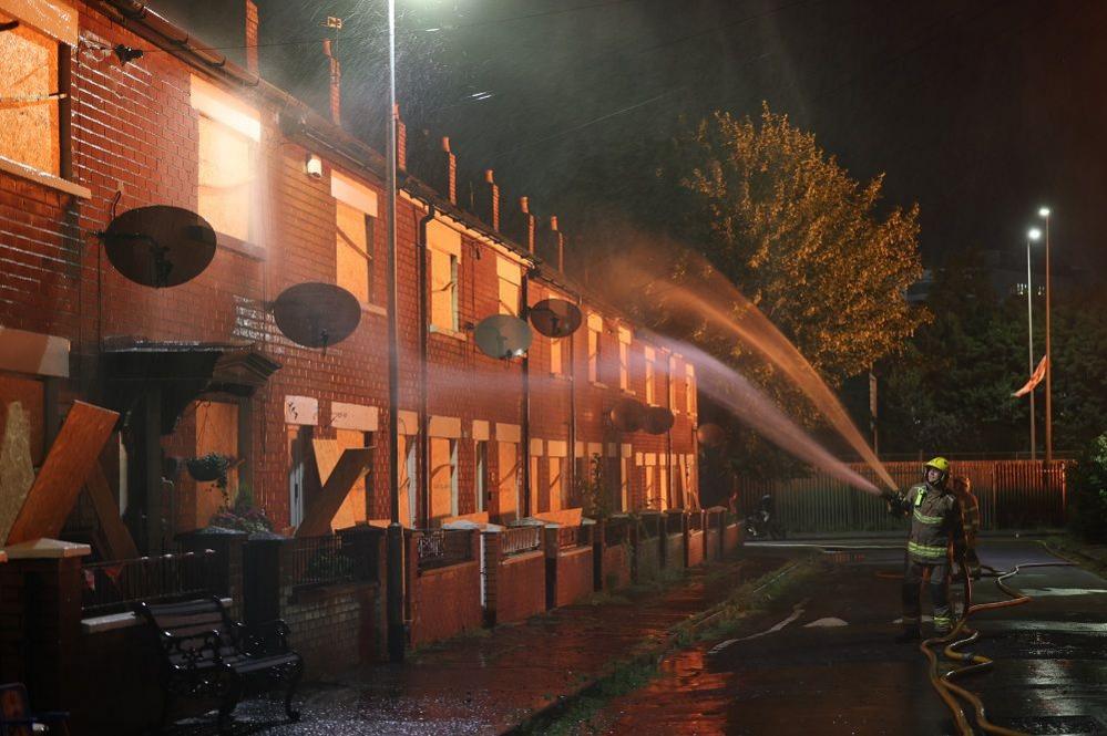 Image of two fire fighters spraying a terraced house with water, the windows are boarded up and it is dark outside, with a red glow from nearby flames