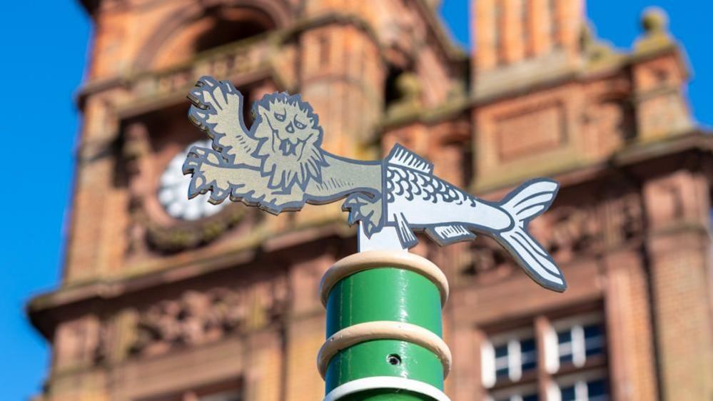 A green post, with a heraldic lions head adjoined to a herring fish tail, which is mounted outside Great Yarmouth Town Hall. The building is built of red sandstone and red brick, with decorative features including friezes and a clock and turret.