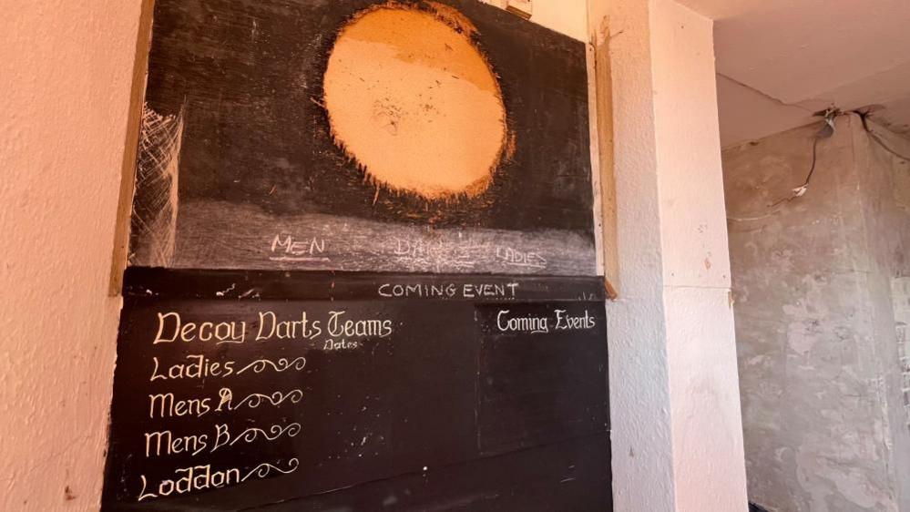 A wall partially painted black with an unpainted disk where a dart board was once hung. The painted words state: Decoy Darts Teams: Ladies' Mens A; Mens B; Loddon and Coming Events.
