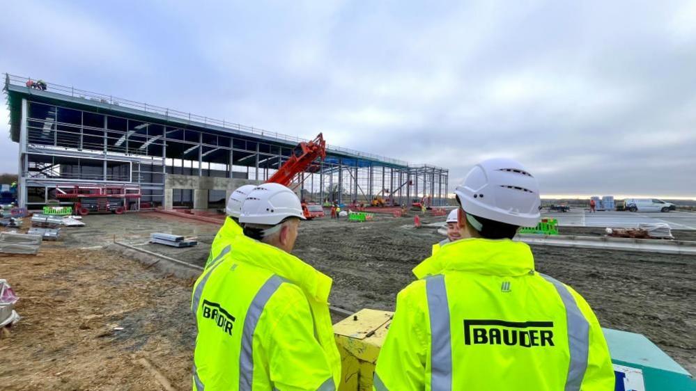 Three men with hard hats and high vis jackets which say "Bauder" on them looking at a warehouse being built. They are standing on a muddy building site. 