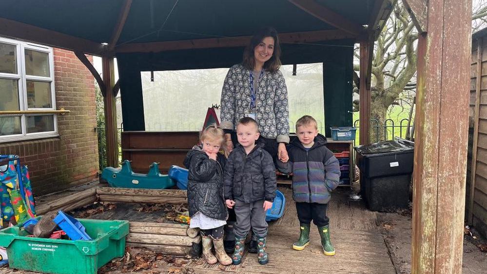 Nursery class teacher Marijka Kirk stands in a large wooden gazebo with three small children all wrapped in thick coats because it is a rainy day. 