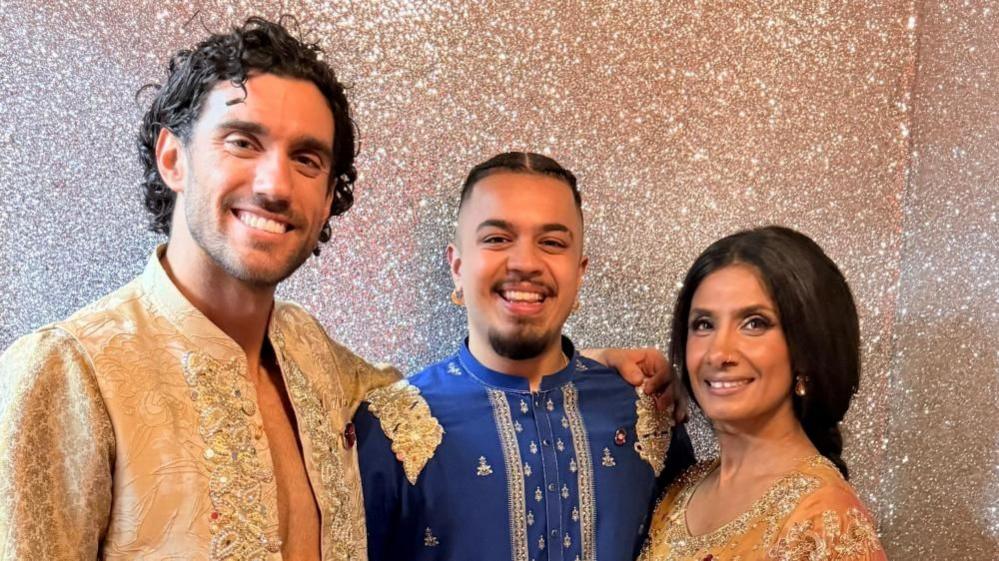 Three people standing in front of a gold glitter wall. The man in the middle is wearing a blue outfit and the man and woman on either side of him are dressed in gold outfits. They are all smiling.