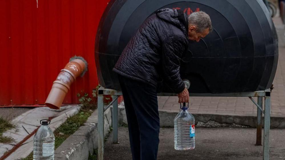 A man fills a plastic bottle with water from a tank located outdoors.