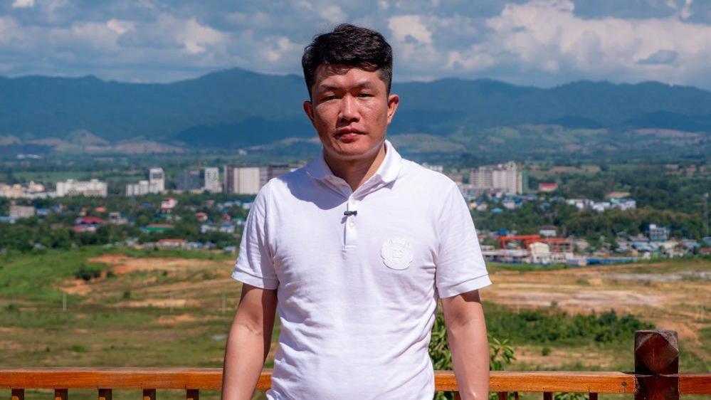 He Yingxiong who is running Shwe Kokko stands in front of a wooden fence with a field and city in the background