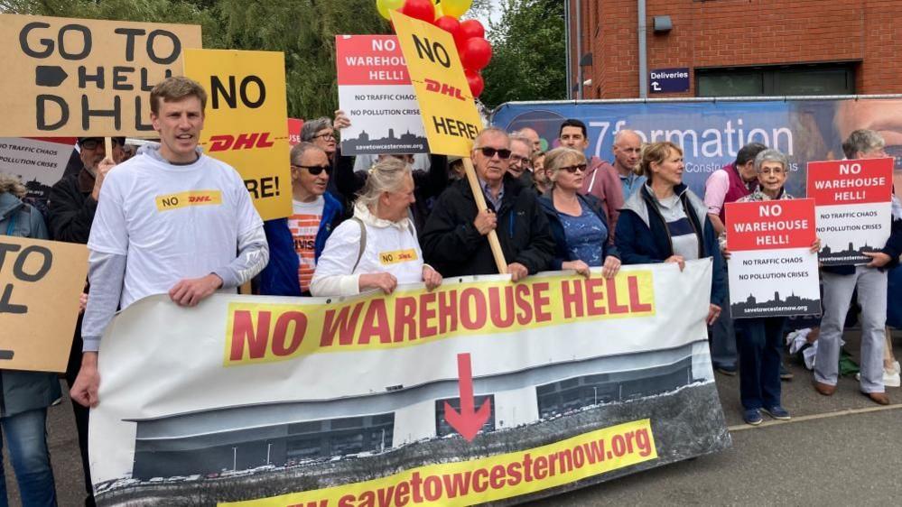 People standing behind a large banner saying "No Warehouse Hell, save Towceseter now". Some are holding placards with messages such as "Go to hell, DHL".