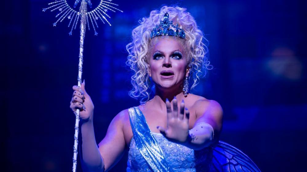 A blond drag queen in a sparkly blue dress holds a big wand and stands with her hand outstretched. 