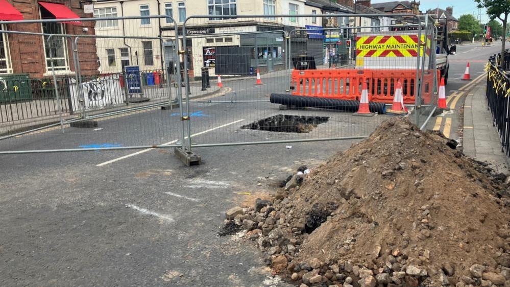 A large pile of dirt and rocks is on a road near a large sinkhole. The hole is surrounded by metal barricades.