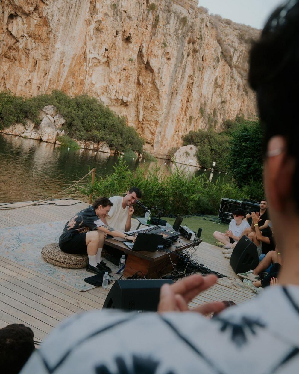 SOAK and Fred Again, again in Greece, performing in front of a cave, sat on the ground with an audience watching