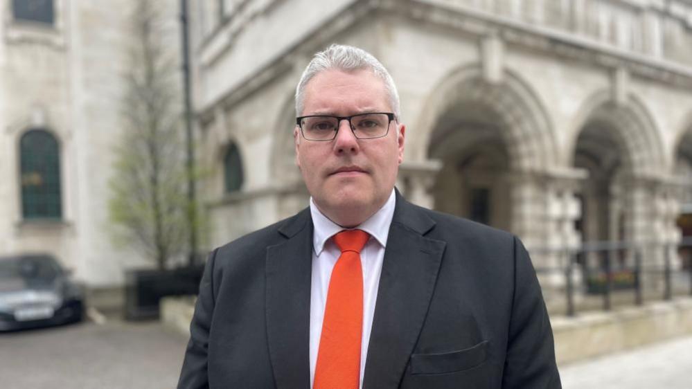 Gavin Robinson with short white hair wearing a red tie