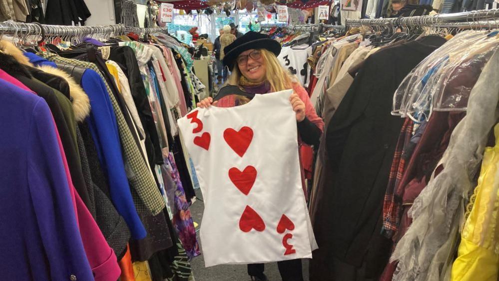 A woman wearing a black hat and glasses holds a top which looks like a three of hearts playing card. She is standing next to racks of clothes