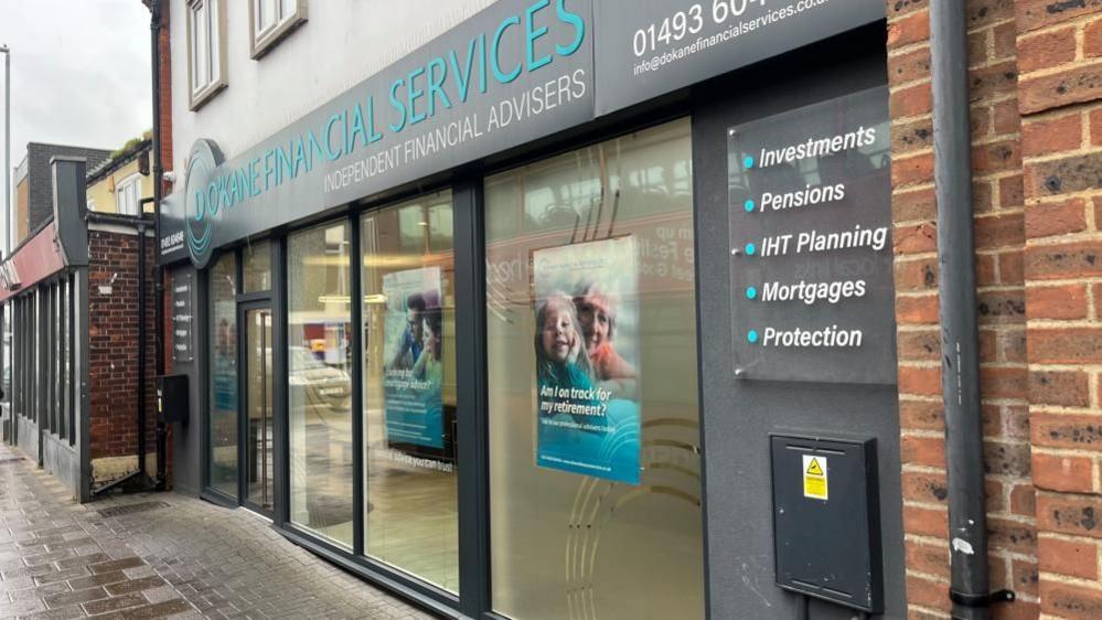 The former NatWest has dark grey window frames, with partially obscured windows. The sign above the windows is in a cyan colour, with white render and white plastic windows on the upper storey