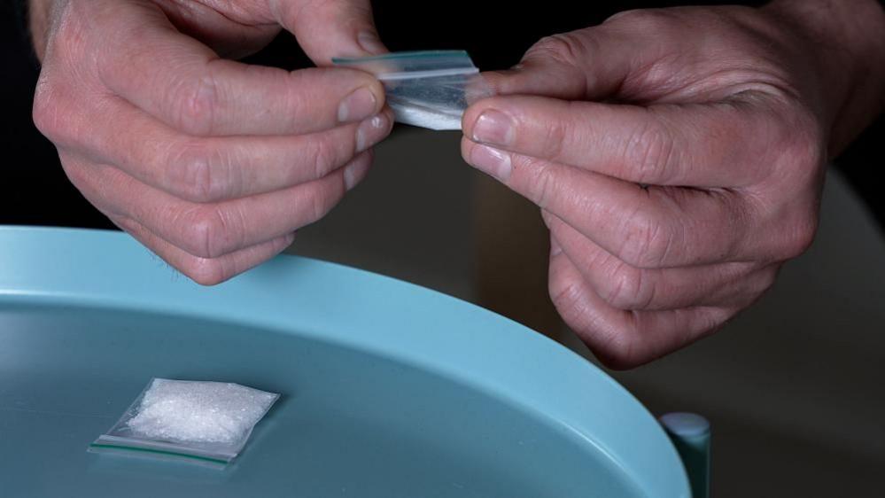 A man's hands are seen holding a small clear bag with white powder inside. A blue table top underneath the man's hands also has a bag with white powder in it.