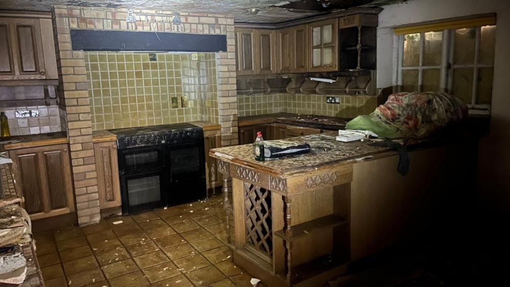 The kitchen in a burned out home, with tiled and brick wals, and counters, covered in debris. Part of the ceiling can be seen with black mould on it.