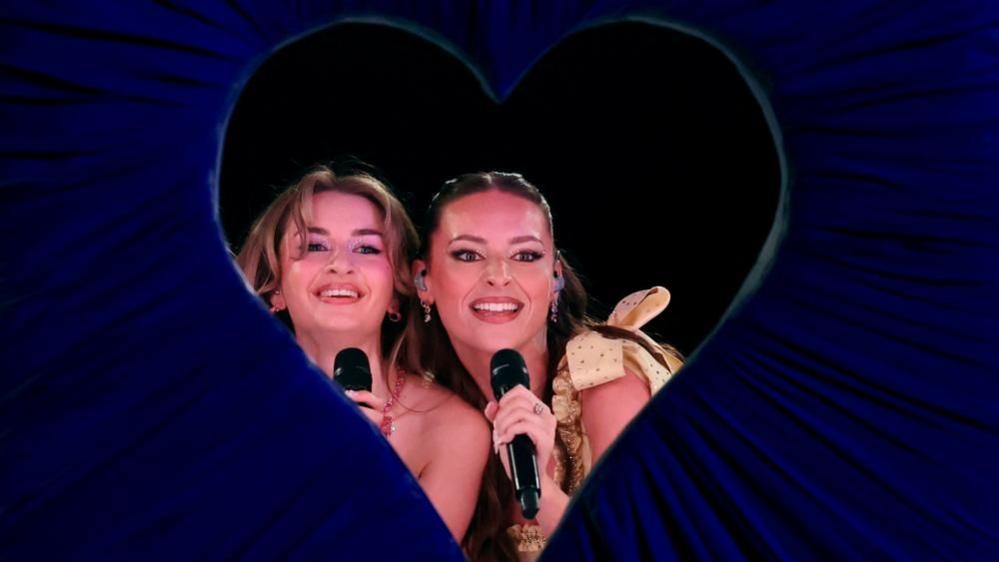 Two of the three members of girl band Remember Monday during a dress rehearsal, looking through a stage prop heart-shaped blue velvet curtain while holding microphones and smiling.