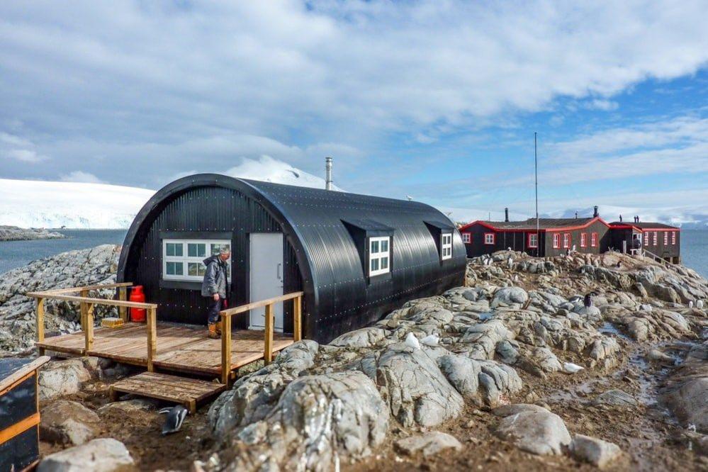 Semi-circle shaped black hut with a small wooden deck, sitting on top of a rocky area with another building in the distance.