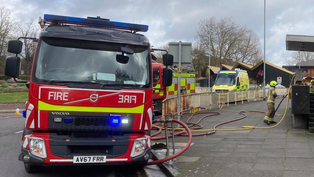 Two fire engines can be seen parked on a road with an ambulance behind. A fire officer with protective gear is on the pavement and fire hoses are too.  