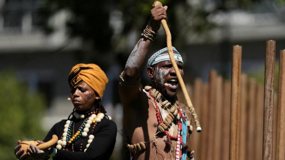 Two praise singers are seen dressed in colourful traditional attire at a ceremony commemorating the military workers. The praise poet in front holds a cane in one hand.