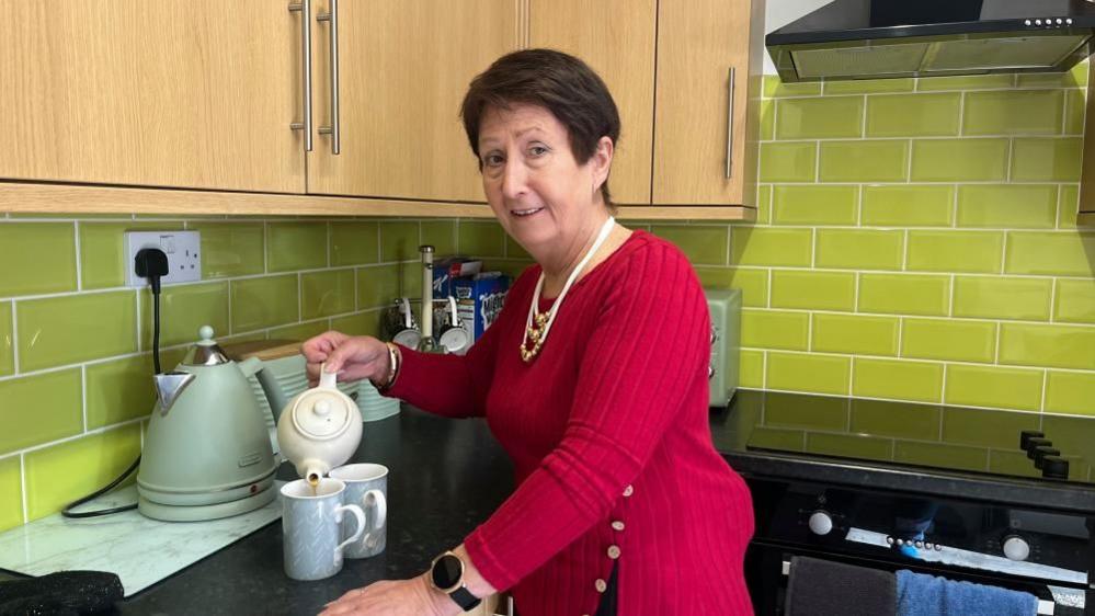 Jan Fuller, wearing a red-knitted jersey which shows a bulge above her left hip where the pump driver is positioned. She is holding a teapot, pouring tea. Nearby is two cups, a kettle and other utensils. There is a set of wood-effect cupboards to the left of the image; the tiled walls are lime green coloured and the kitchen worktop is black.