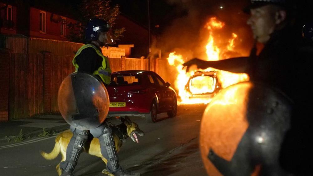 A police officer in riot shield with a dog walks in front of a burning car during a riot.