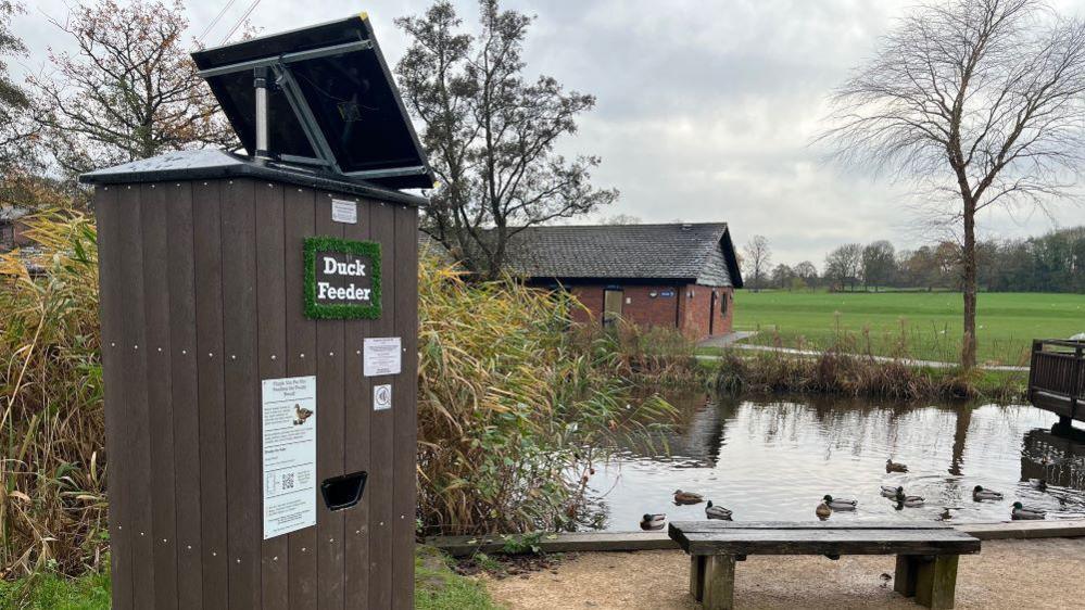 In the foreground is a brown wood-effect box with a hole for dispensing bird feed. There is a bench nearby, and in the background is a pond with a number of waterfowl resting on the surface. Further in the distance is an open field surrounded by trees.