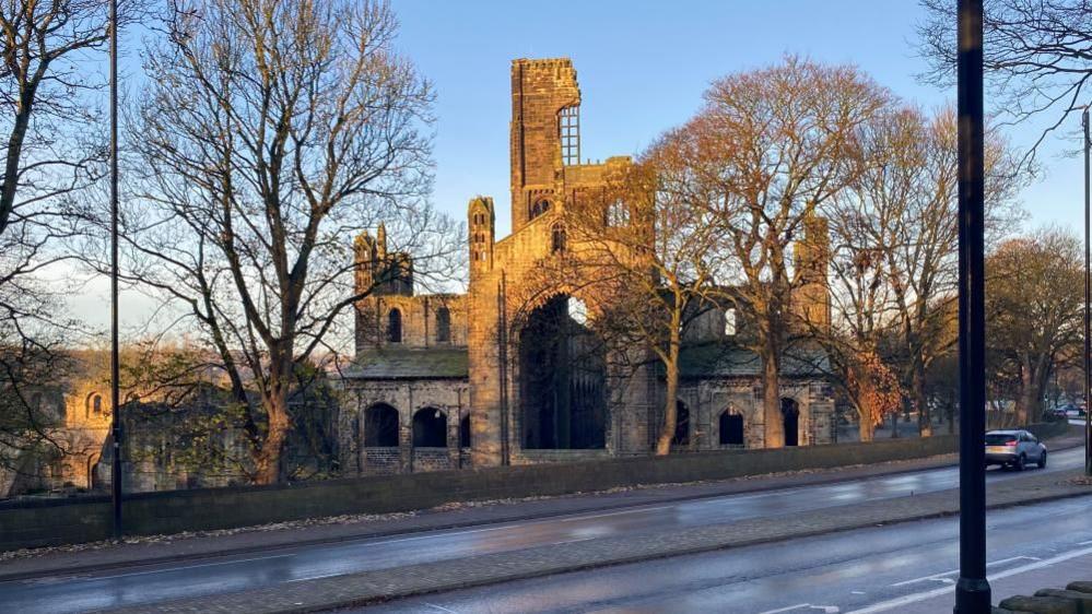 A ruined abbey on a cold winter's day. It is surrounded by trees and viewed from the opposite side of a main road.