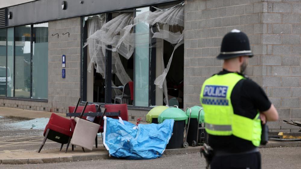 Broken windows and chairs outside the Holiday Inn Express at Manvers, with a police officer standing nearby