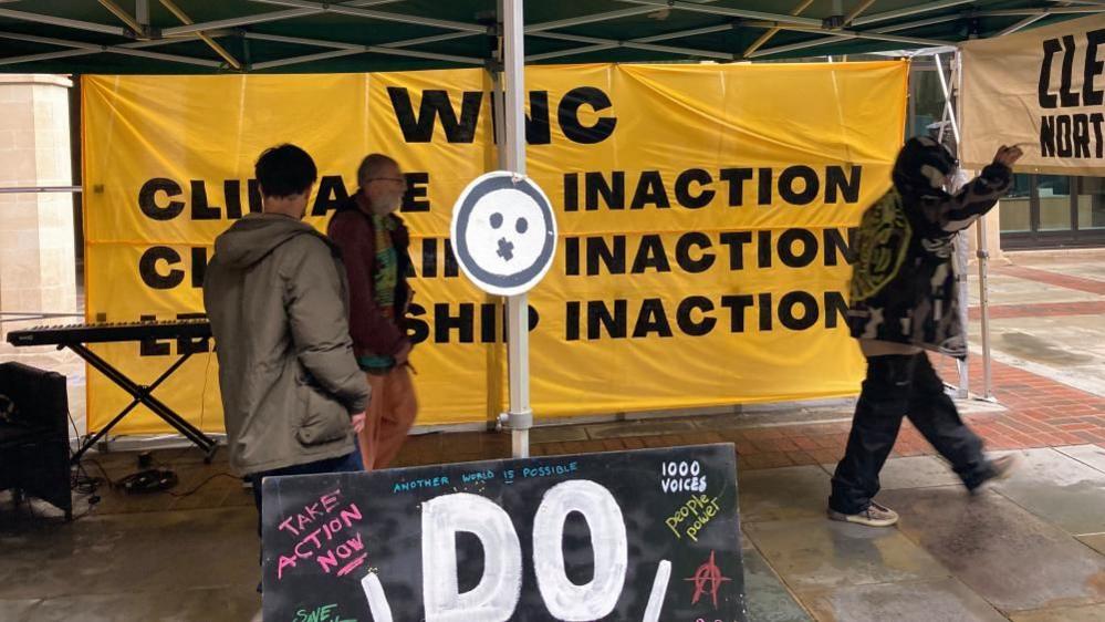 Large yellow banner with the words "WNC Climate Inaction" repeated several times. It is inside a gazebo. Three people are standing beside it. There is also a black placard with 1,000 Voices and various other slogans.