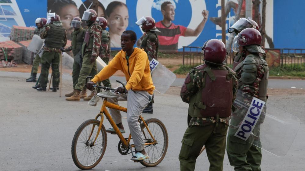A cyclist passes a line of riot police in protective gear blocking a road in Nairobi 