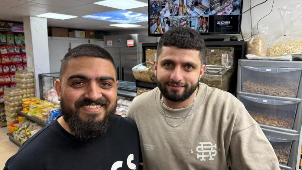 Two men, one in a dark top and one in a lighter top, are standing side by side and smiling. They are in a shop and there are a variety of items in the background.