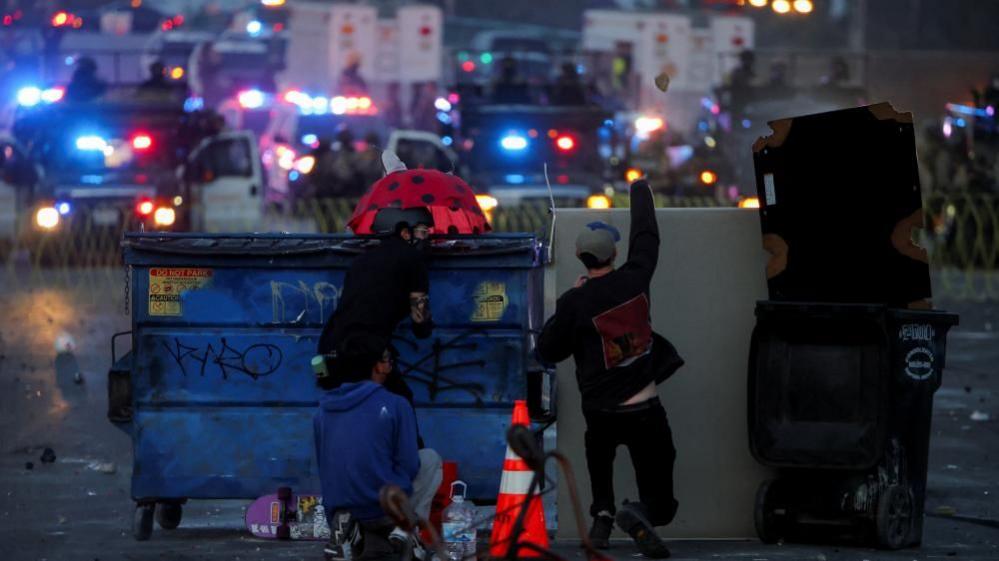 Two men crouch behind large bins with riot police in the distance