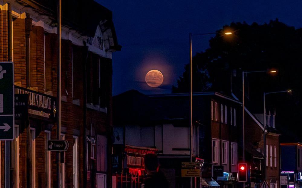 A pink Moon is seen in the sky over the roofs of terraced homes and businesses in a town. It is night-time and lamp-posts are illuminated.