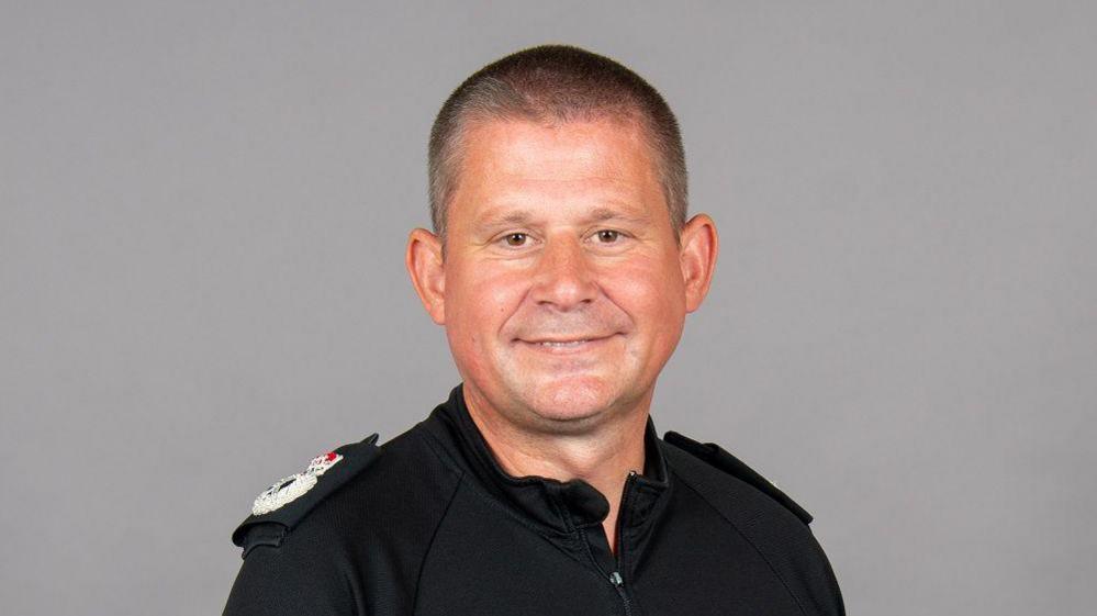 A man in a black police uniform looks directly into the camera smiling. 