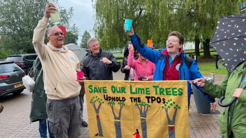 Campaigners with their arms aloft behind a banner saying "Save our lime trees - uphold the TPOs (tree preservation orders)". The banner also has a picture of four trees and a woman in a pink blouse standing between them. Some trees are visible behind the campaigners.
