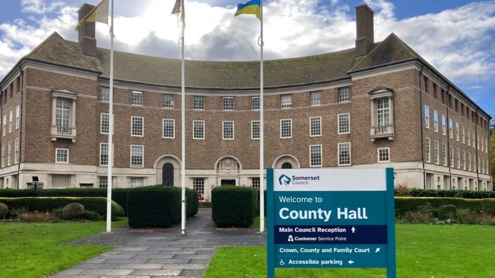 Somerset County Hall is shown in a mix of sunshine and cloud, with a Ukrainian flag visible on a flag pole in the foreground. The ground is slightly damp, presumably from earlier rain.