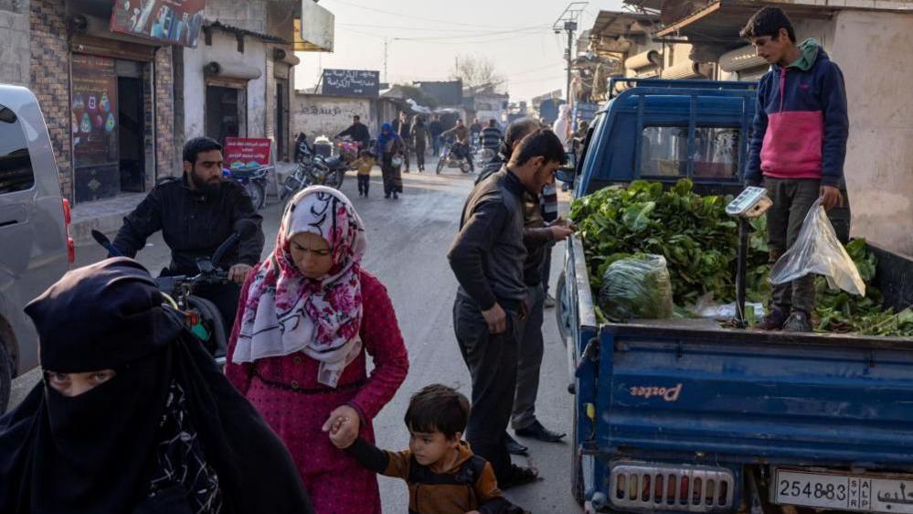 People walk along a busy street at a refugee camp in Idlib with several people buying vegetables from the back of a truck on 17 December