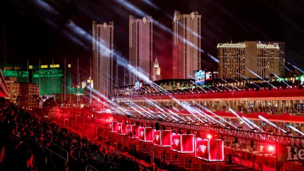 Stands at the F1 Grand Prix in Las Vegas. Streams of light are seen coming from one storey of the stands. There are lighted boxes around a lower storey, and three towers are visible in the background.