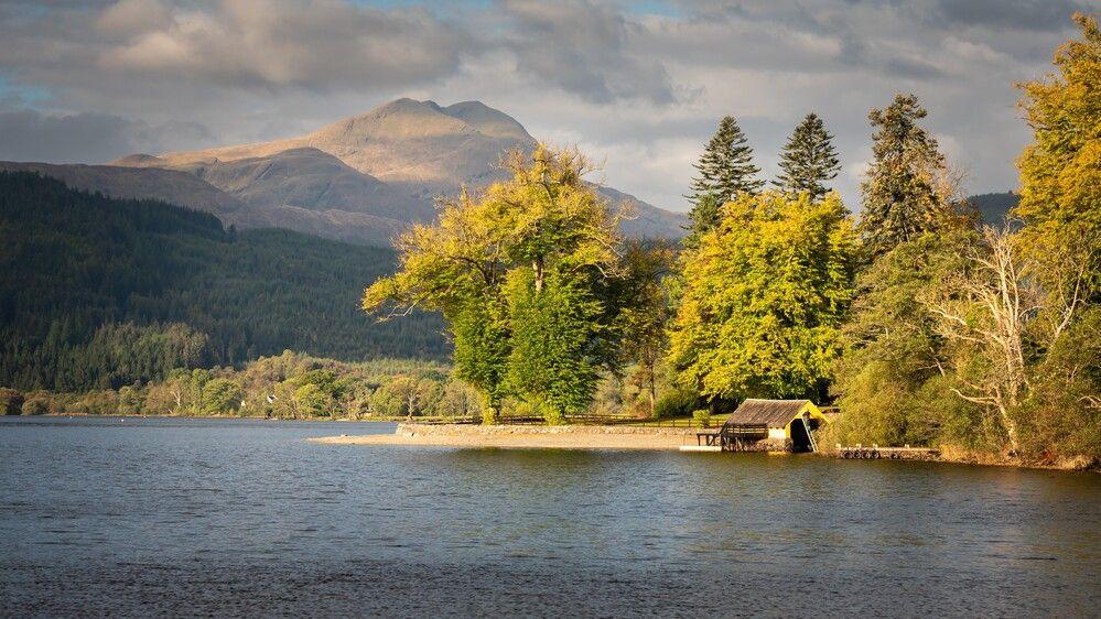 The water of Loch Ard is in the foreground and in the distance there's a beach with a little boathouse. Large trees are behind the house, with a forest in the background and a tall munro is behind the forest.