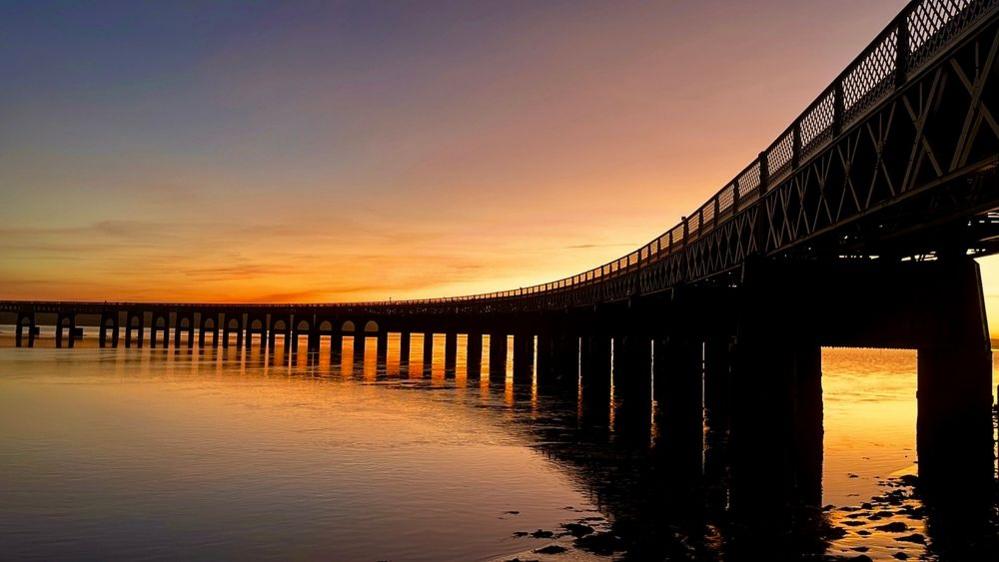 The Dundee Tay Rail Bridge curves across the River Tay. It's black against a sunset sky which reflects on the water.