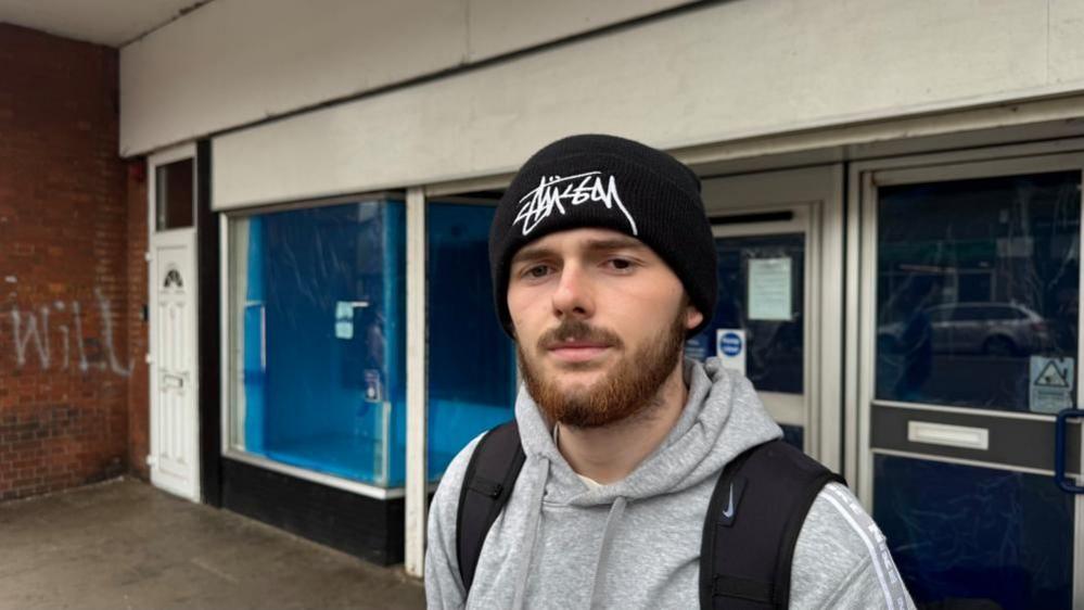 Harry Barnard is wearing a grey hoodie and black knitted hat, and black rucksack with wide straps over his shoulders. He has a full dark/ginger beard and moustache.