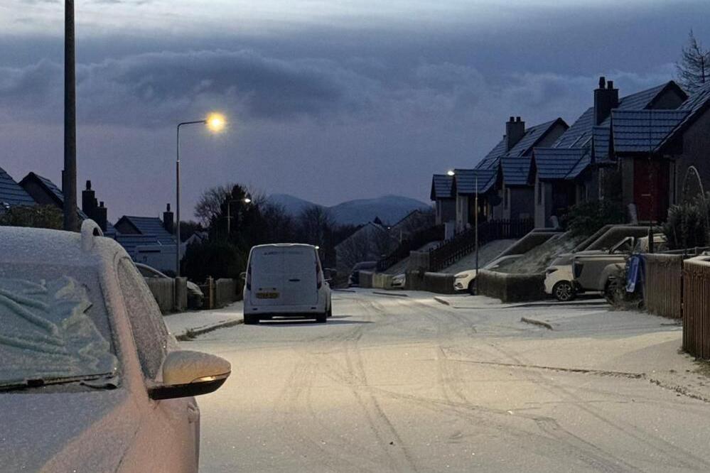 A street with houses on both sides and cars parked along the road. The road has a light dusting of snow with a car covered in ice nearby.