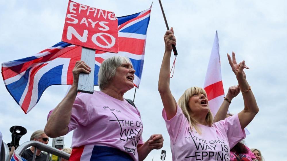 Protesters attend an anti-immigration demonstration outside The Bell Hotel in Epping. An older woman is wearing a pink t-shirt and holding a placard which says "Epping says No". There are Union and St George flags in the background.