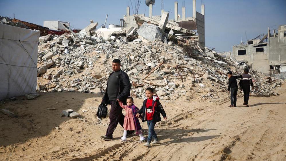 Palestinian man with two young girls walking past the rubble of buildings destroyed in previous Israeli strikes, as two boys walk away in the opposite direction, in Khan Younis in the southern Gaza Strip on 16 January, 2025
