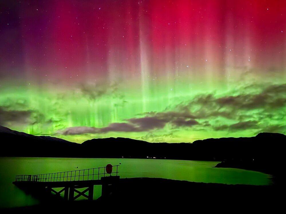 The pink and green lights of the Aurora borealis over the water at Glenelg. The hills make black shapes under the intense lights. 