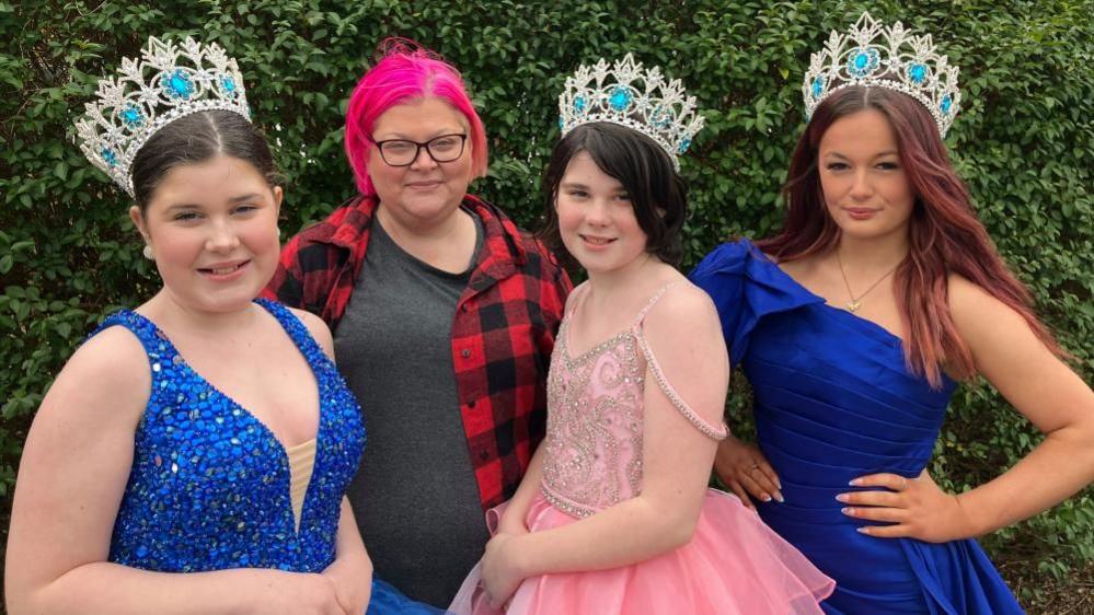 Three girls wearing ball gowns and sparkly crowns stand outside smiling beside a woman with bright pink hair. 