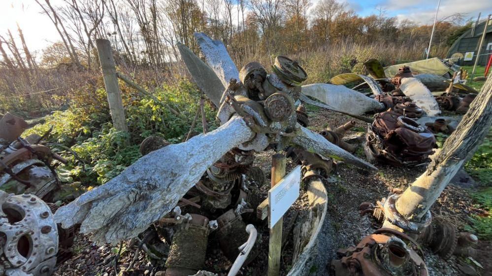 The engine, with its three propellers, is lying on the museum ground surrounded by other piece of planes and trees.