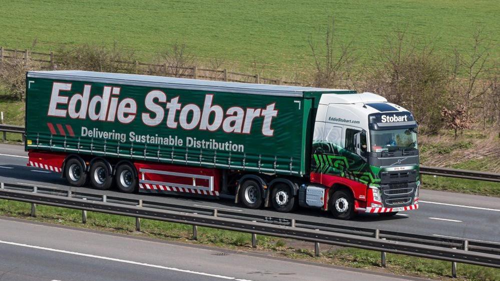 A green, white and red truck driving on dual carriageway. It has 'Eddie Stobart Delivering Sustainable Distribution' in white writing. A field can be seen to the side of the road.