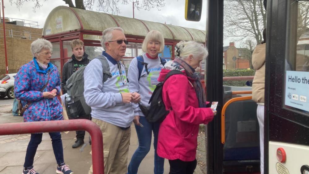 Nick and Kerstin wearing grey sweatshirts and backpacks standing in a queue for a bus, which has arrived. A woman in a pink coat is ahead of them and about to step on to the vehicle. There are two other passengers behind Mr and Mrs Banham. A bus shelter with glass and red frames is behind them.